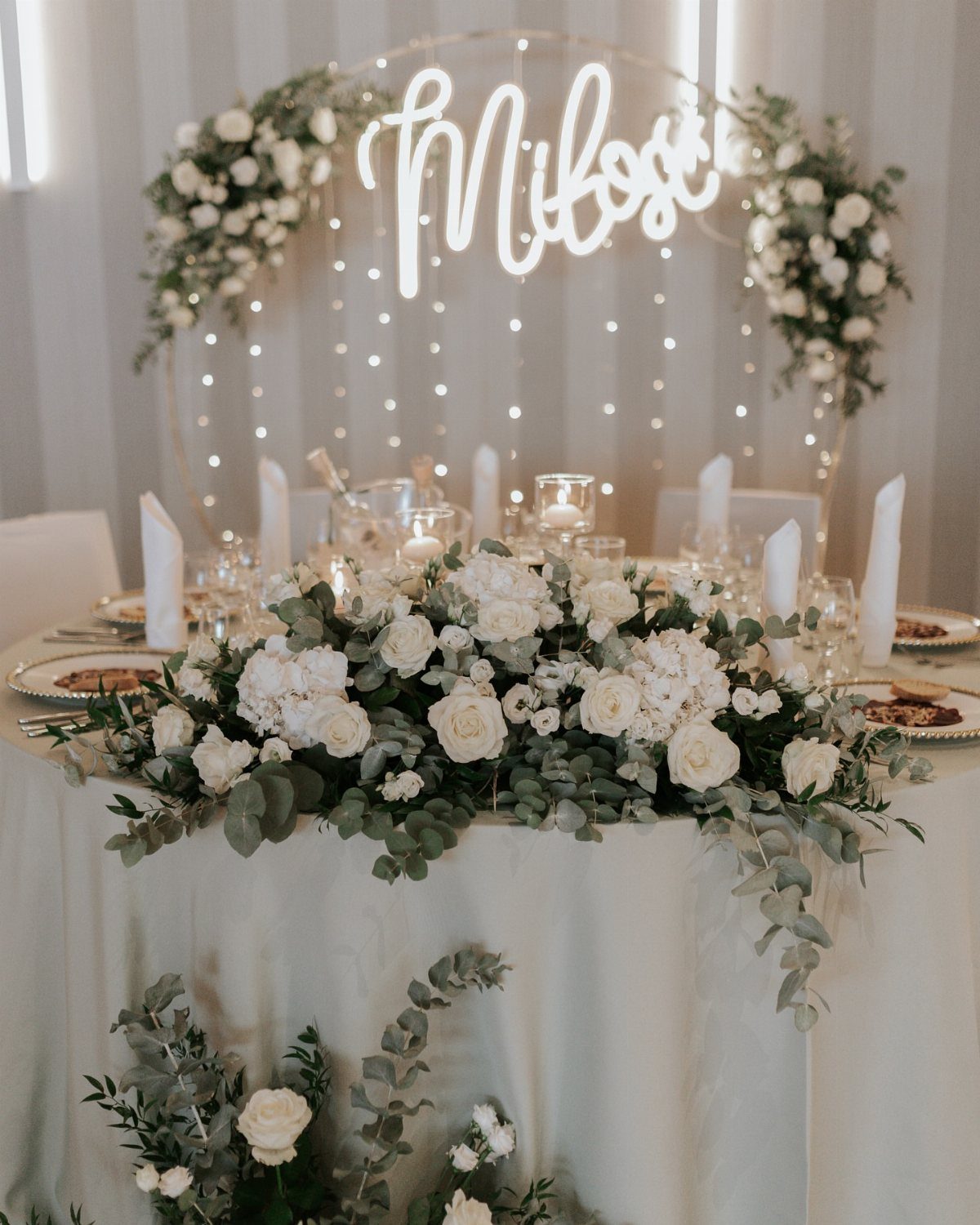 White roses and lush greenery centerpiece on a wedding table with neon sign.