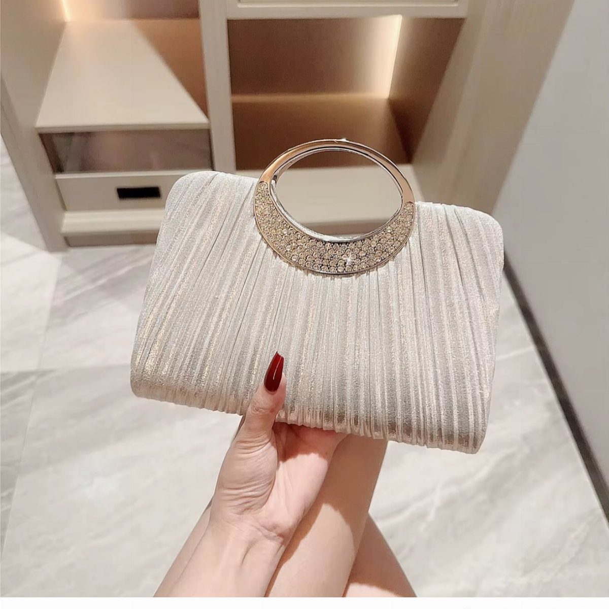Champagne pleated clutch bag with a crystal-embellished circular gold handle held by a hand.