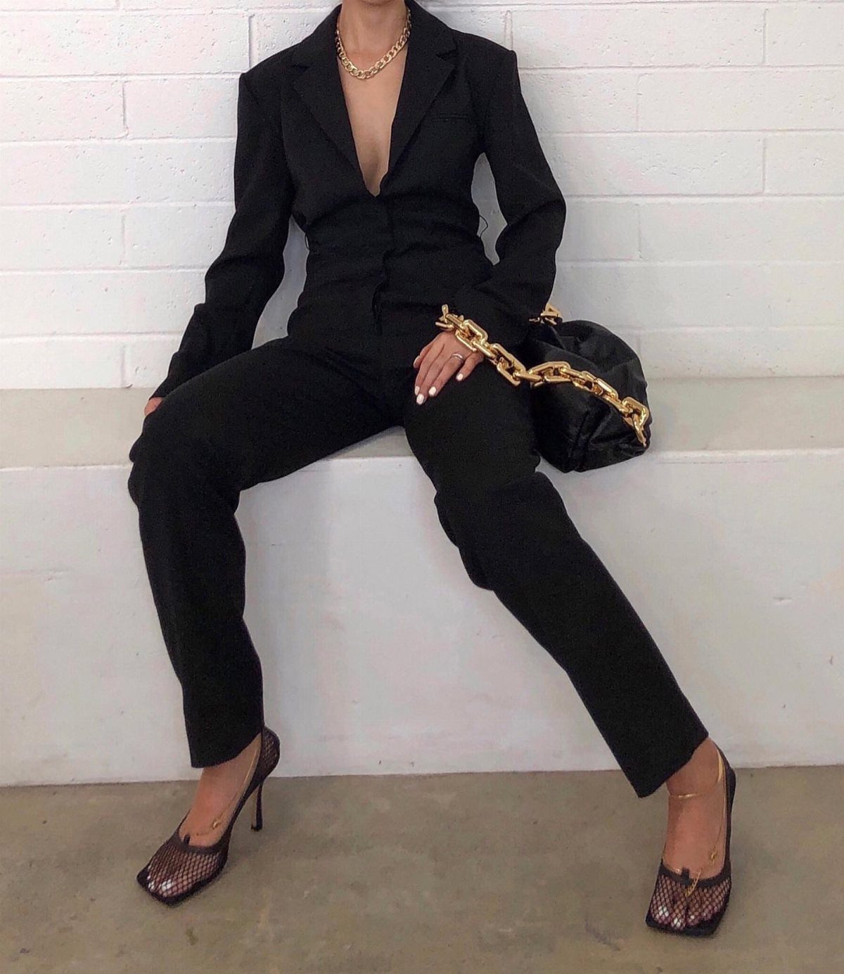 Person in a tailored black plunging-neckline jumpsuit with patterned heels and gold chain bag.