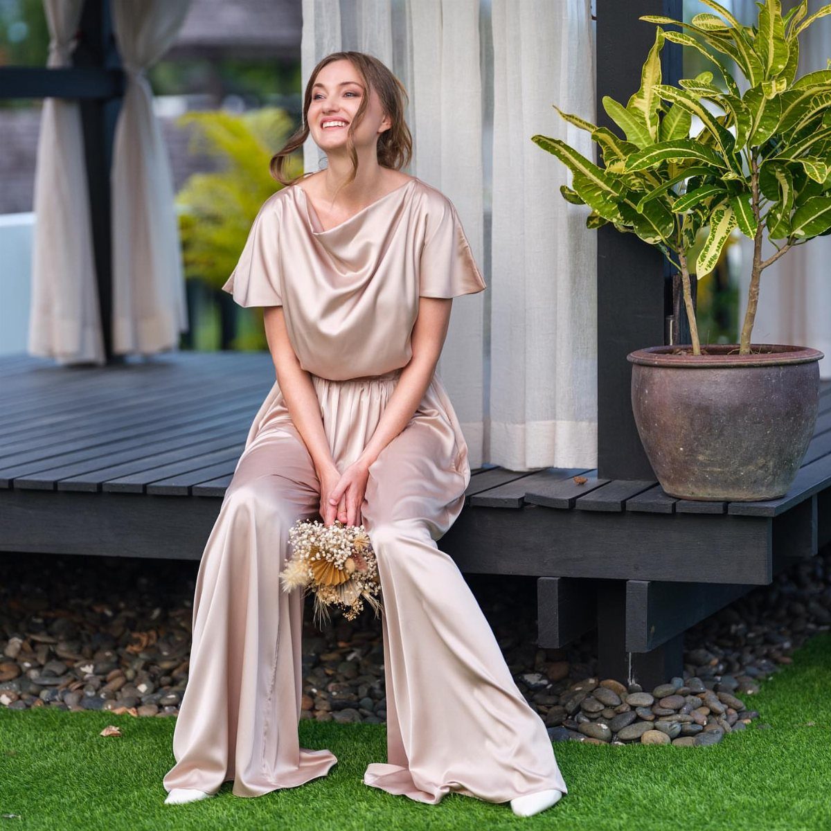 Woman in a champagne satin wide-leg jumpsuit sitting on a wooden deck holding flowers.