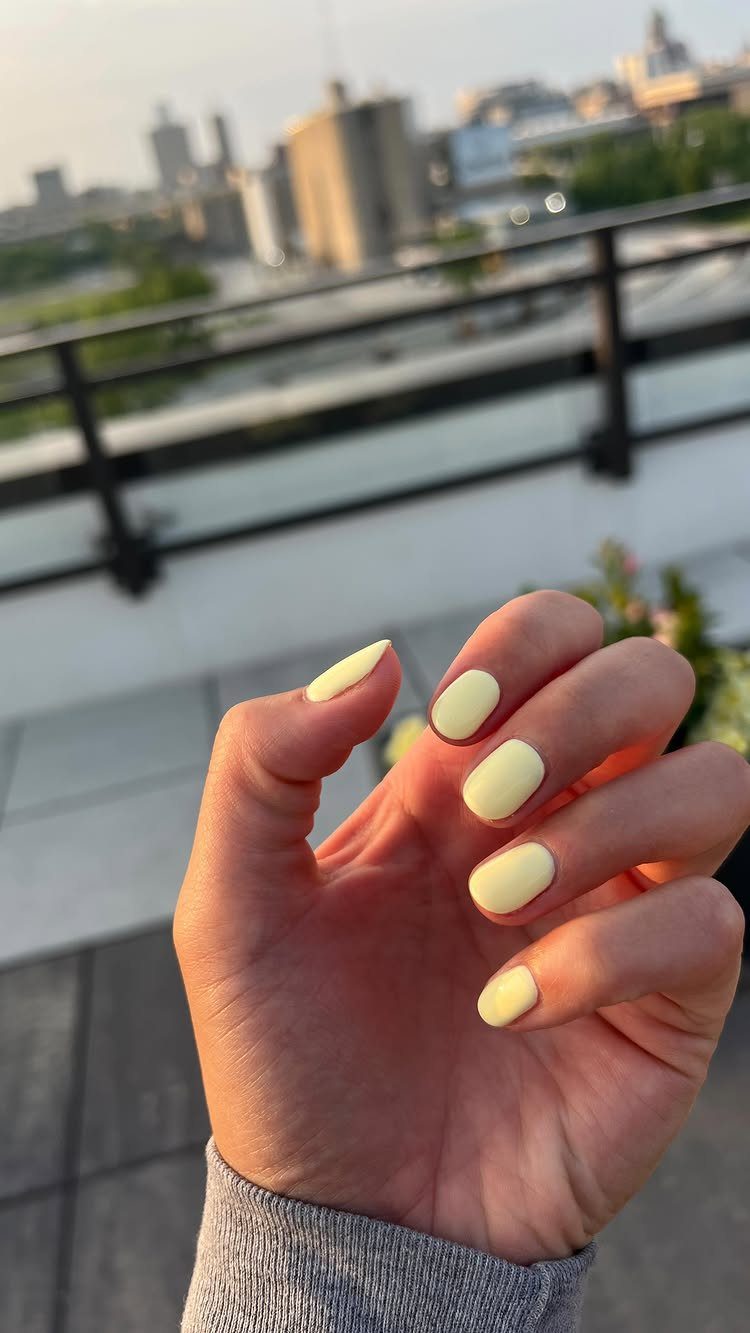 Hand with soft pastel butter-yellow nails posed against a blurred city balcony background.