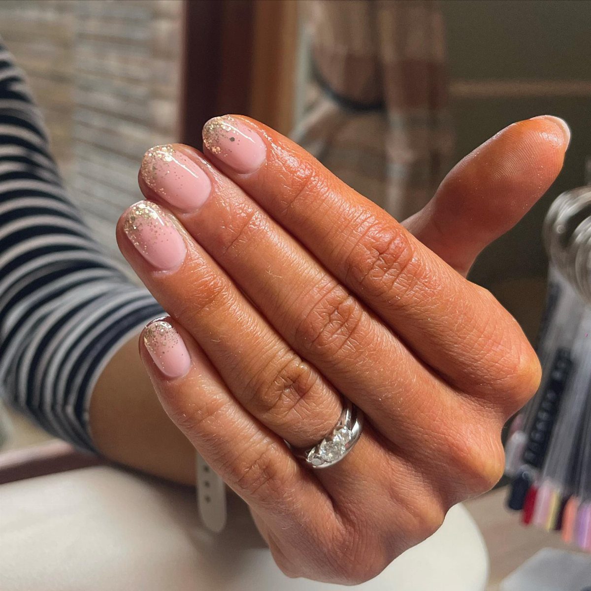 Nude pink almond nails with gold glitter accents on a person's hand.