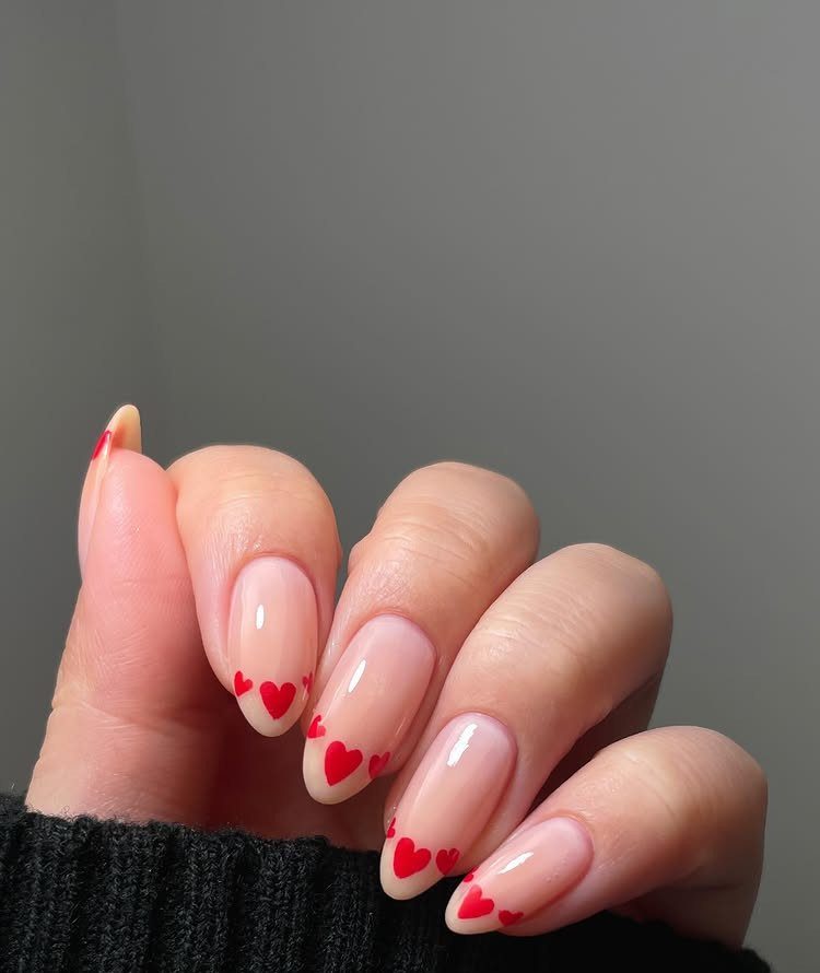 Nude oval nails with small red hearts painted at the base of each nail.