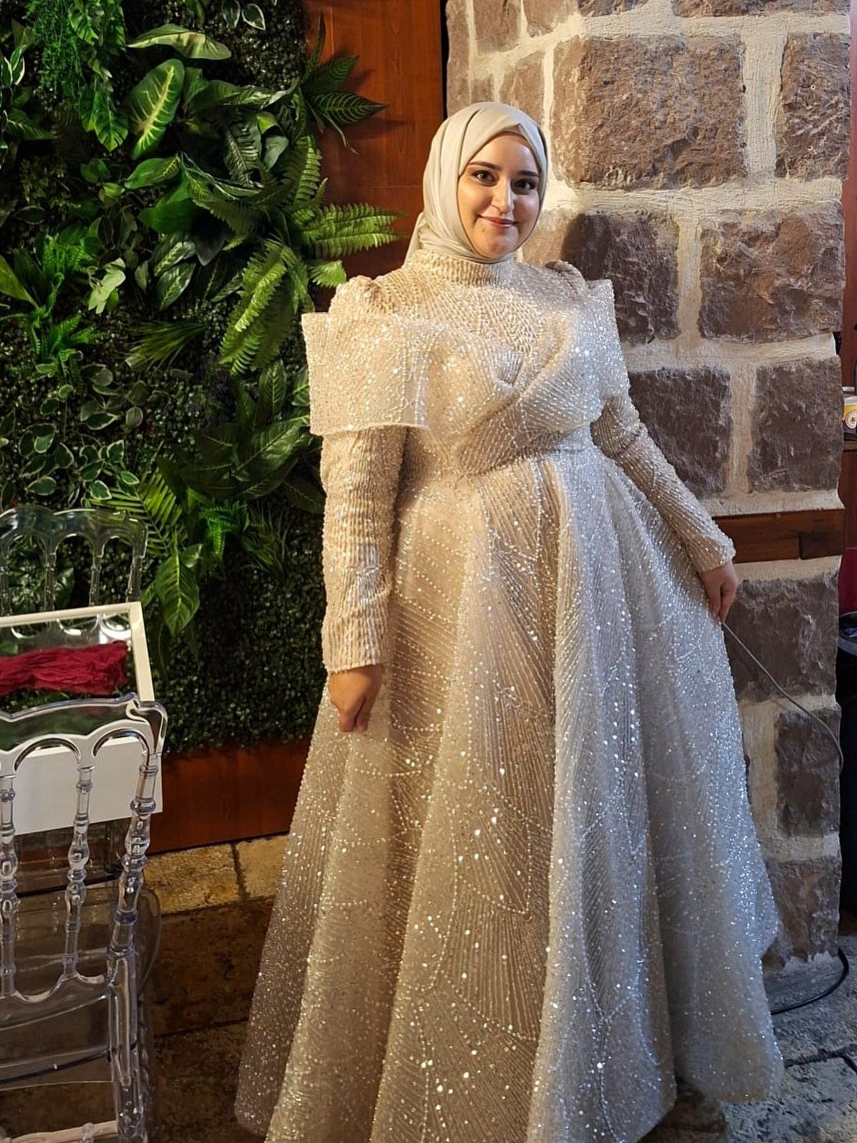 Woman in a shimmering champagne gown and hijab standing by a green wall.