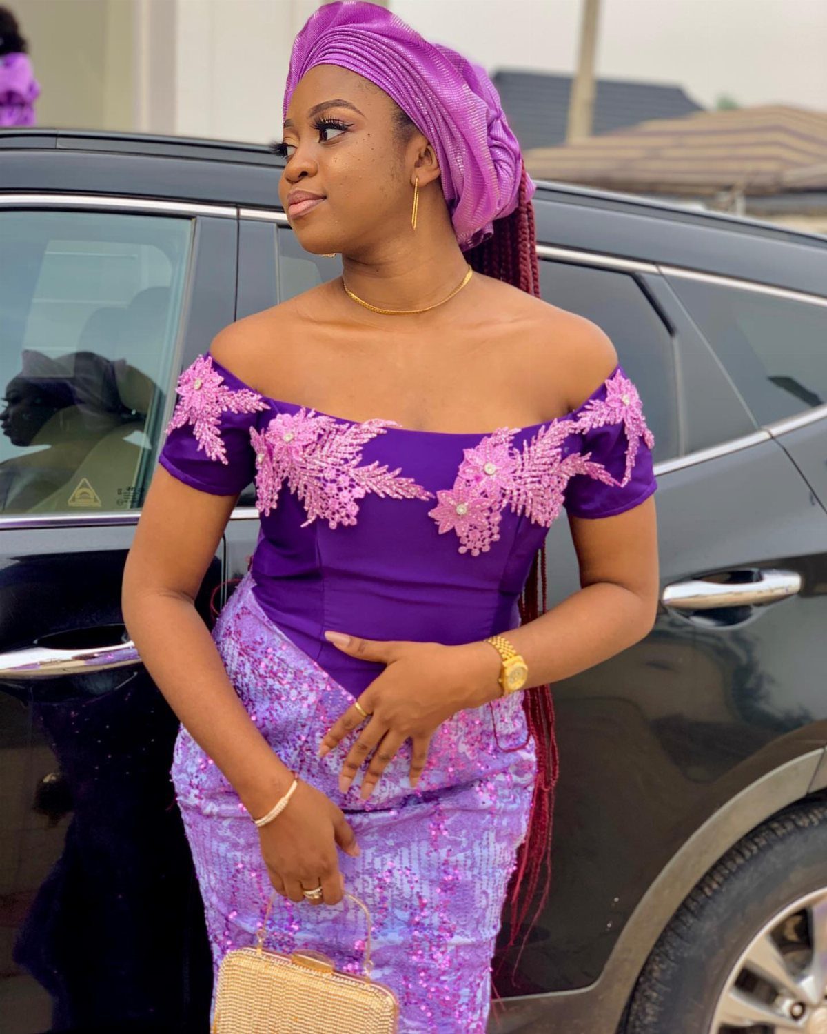 Woman in purple off-shoulder gown and matching gele head tie standing by black car