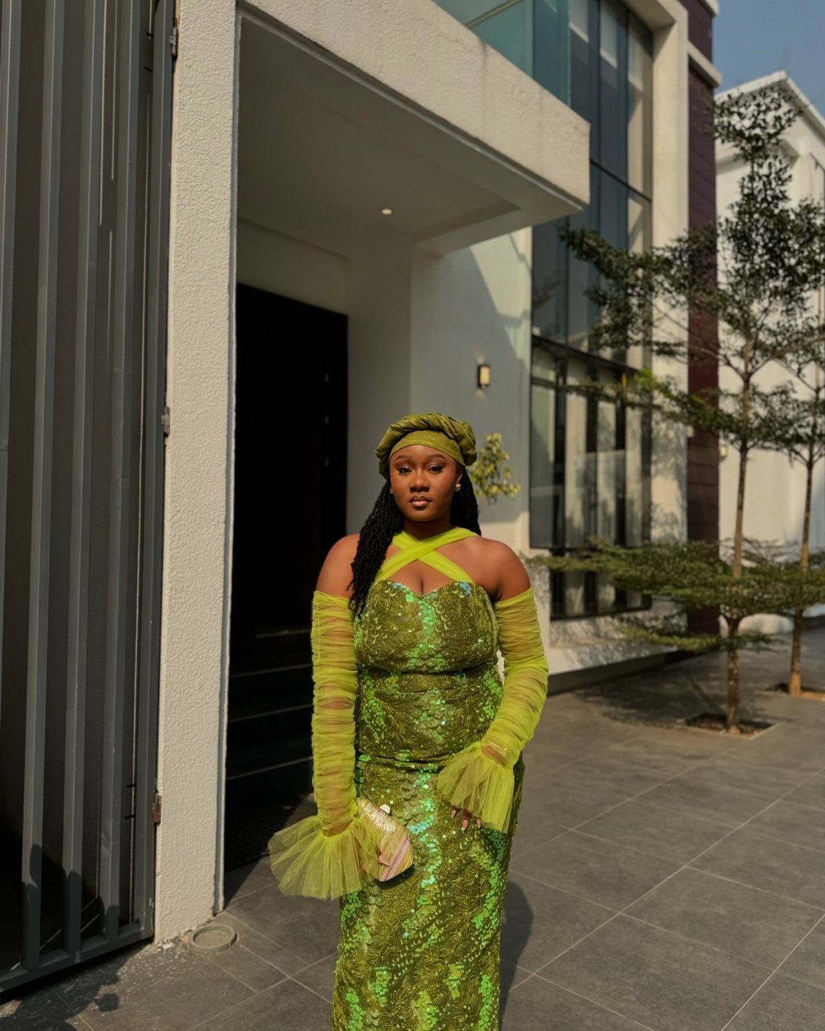 Woman in a lime green sequined gown and headwrap posing by a modern building.