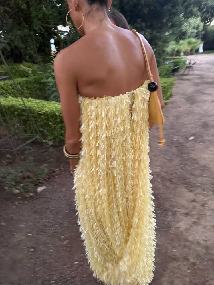 Woman from behind in a strapless yellow fringed dress walking on a garden path