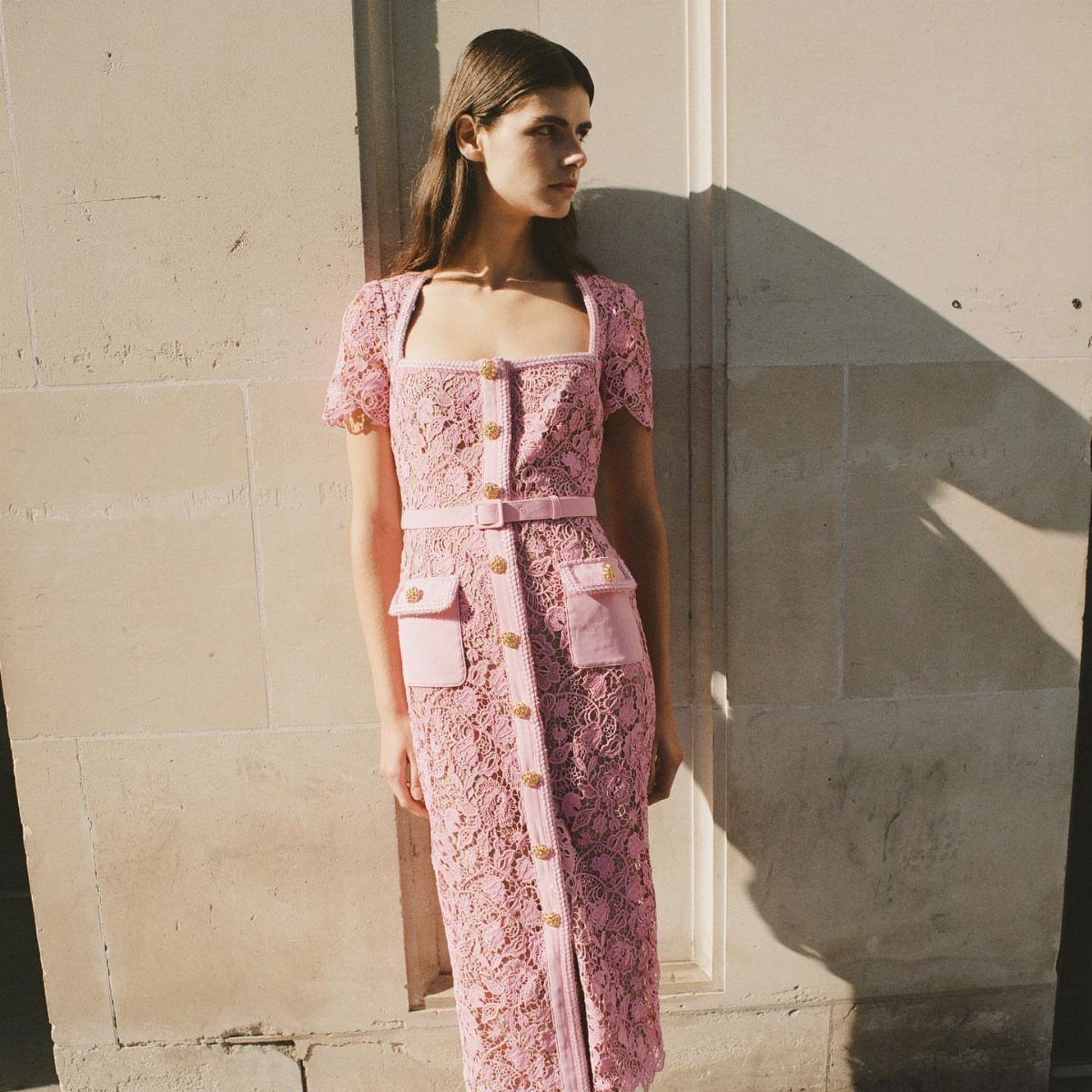Woman wearing a pink lace midi dress with gold buttons and a matching belt.