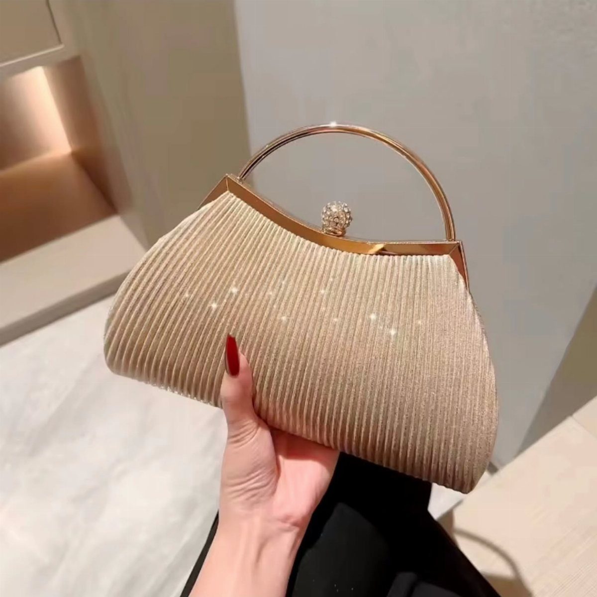 Champagne pleated clutch bag with gold handle held by a hand with red nails.