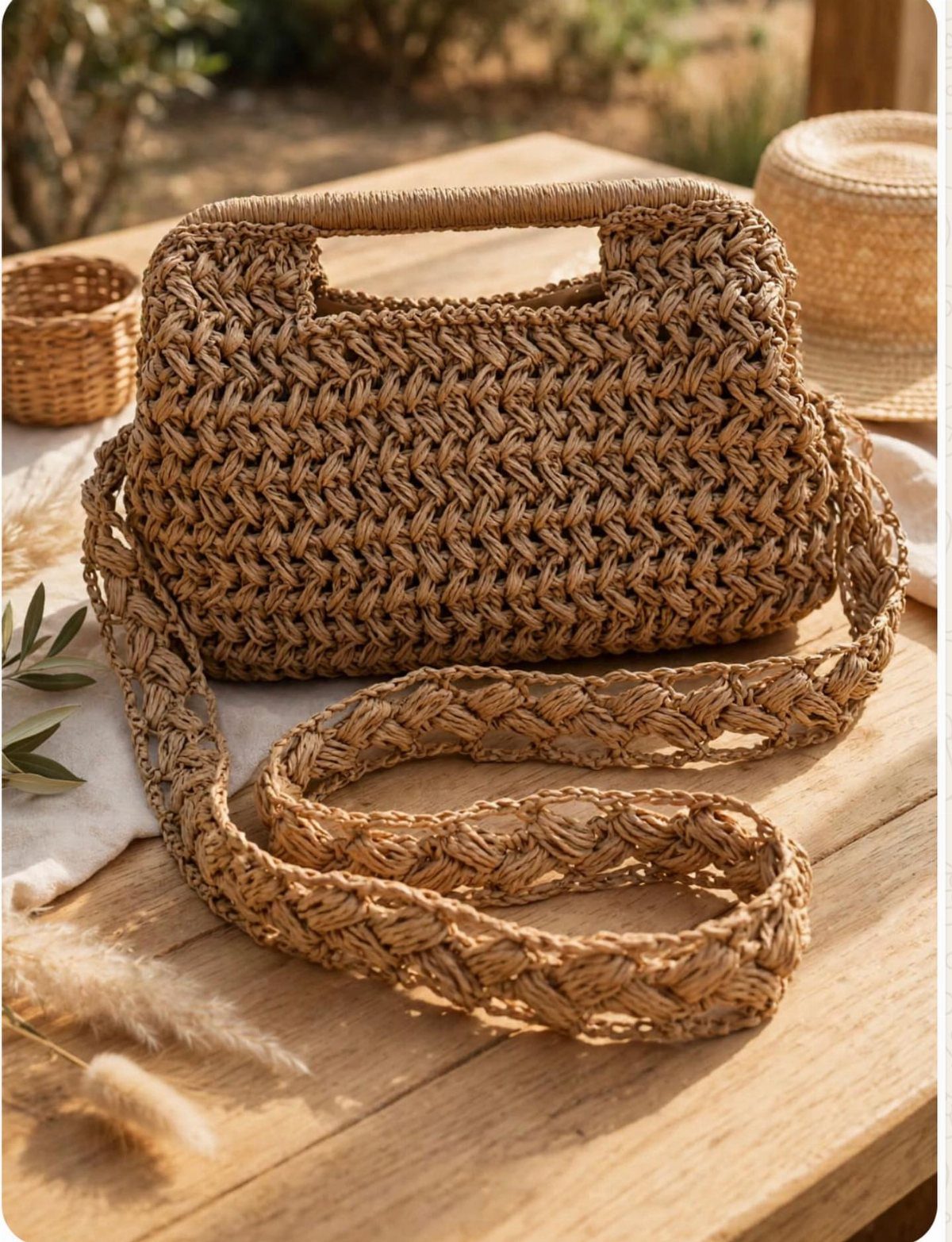 Woven raffia handbag with a top handle and shoulder strap on a wooden table.