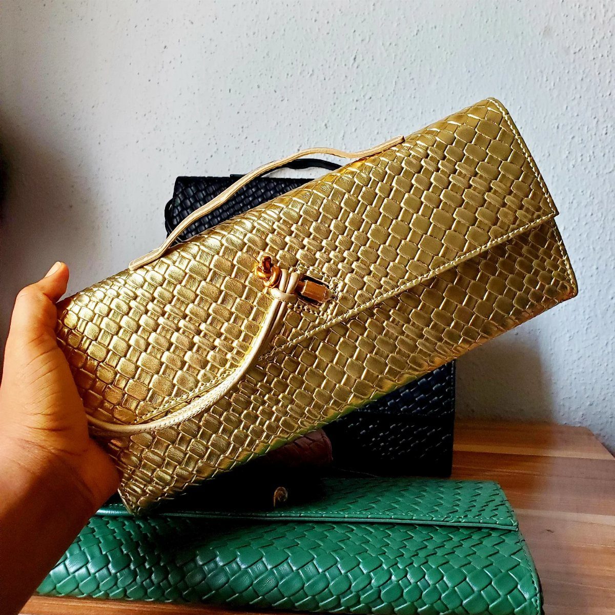 Gold woven envelope clutch bag held by a hand over other colorful bags.