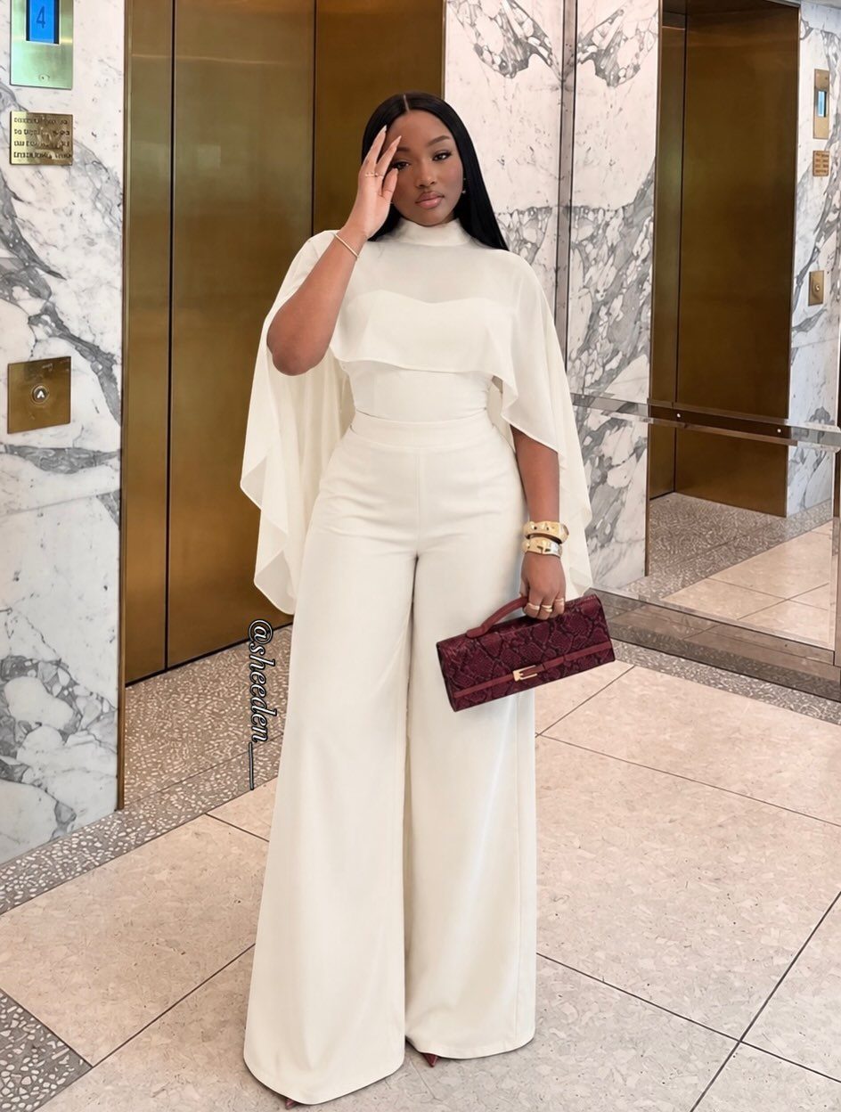 Woman in a white wide-leg jumpsuit with cape overlay and burgundy clutch bag.