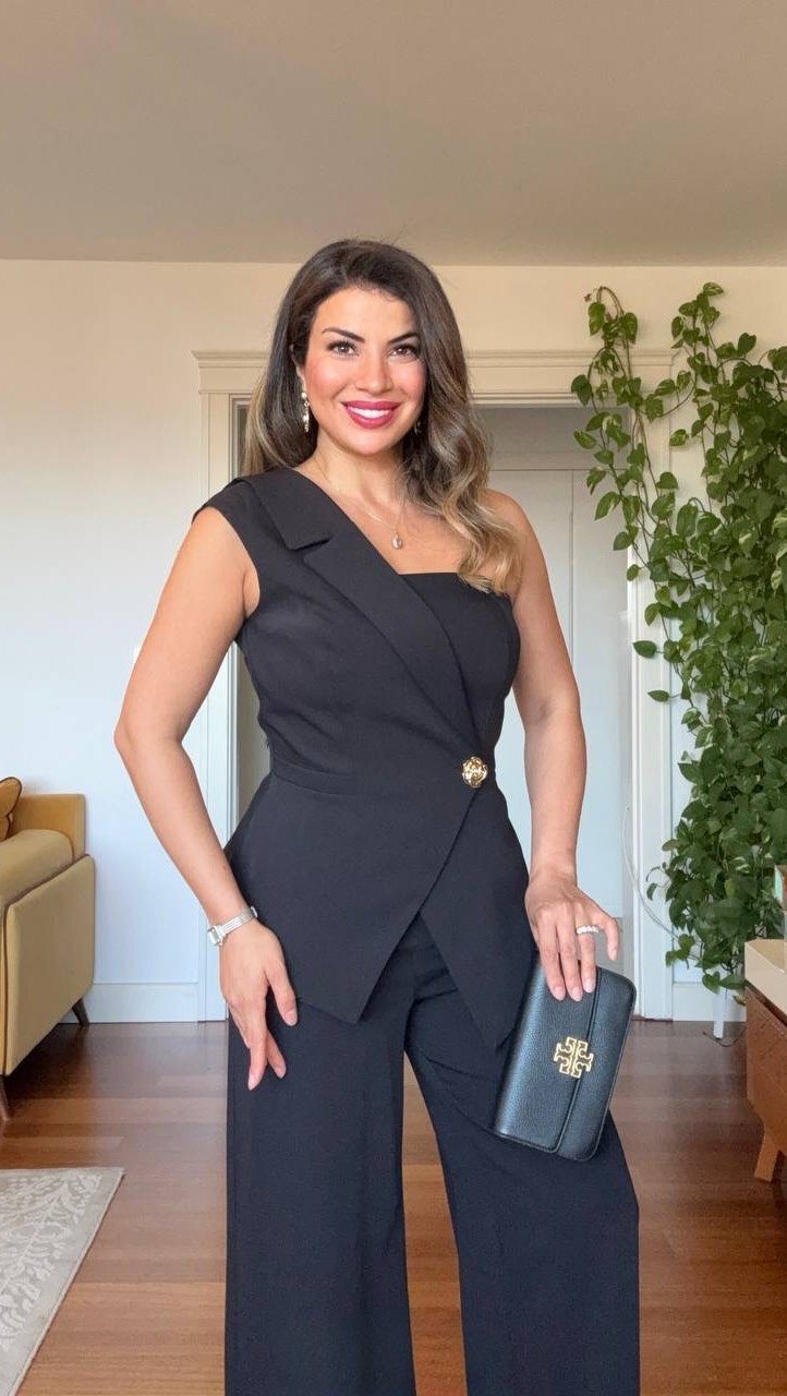 Woman in a black one-shoulder wide-leg jumpsuit holding a black clutch bag.