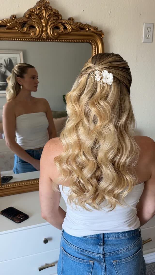 Woman with blonde wavy hair and floral accessory posing before a gold mirror