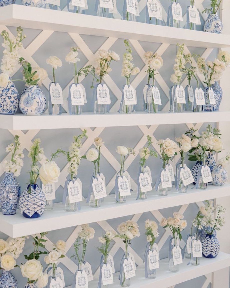White flowers in small bottles and blue vases with attached name tags on shelves.