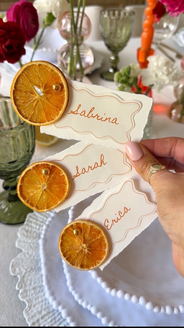 Hand holding white place cards decorated with dried orange slices on a wedding table.