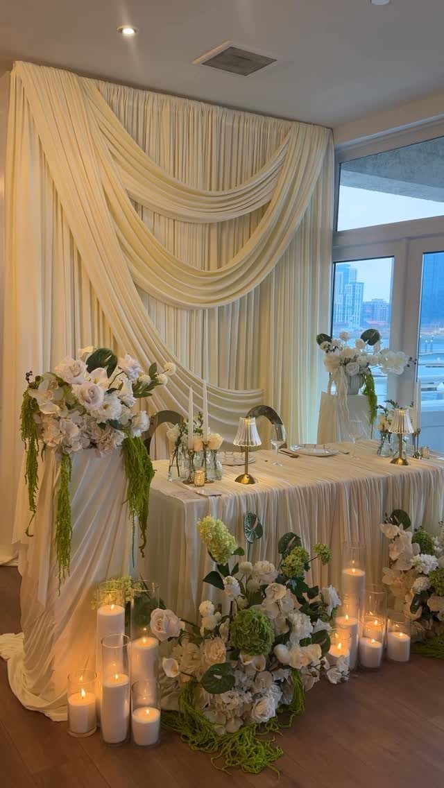 Small white wedding table with floral arrangements and candles against a draped backdrop.
