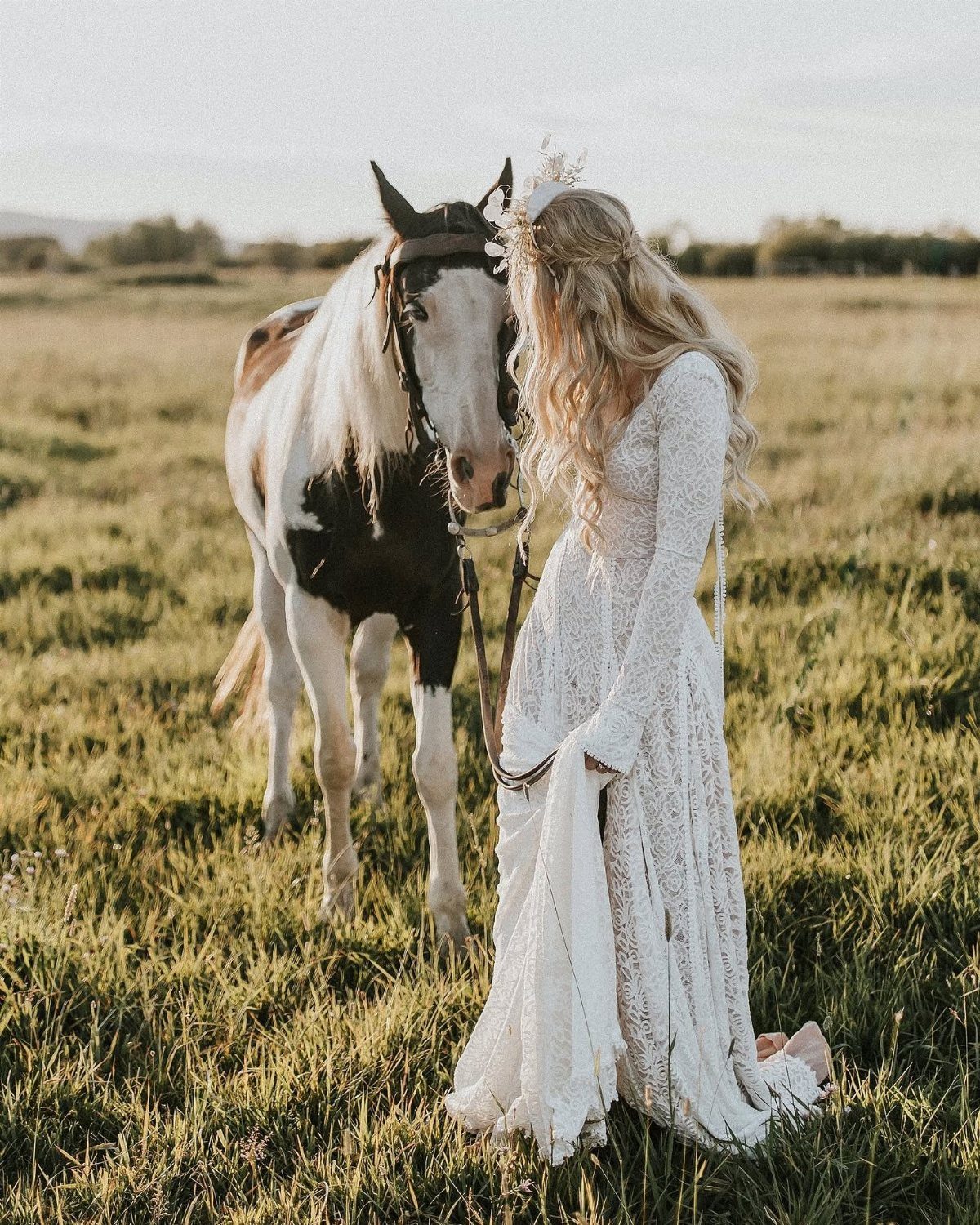 Woman in lace wedding dress standing with a horse in a grassy field.