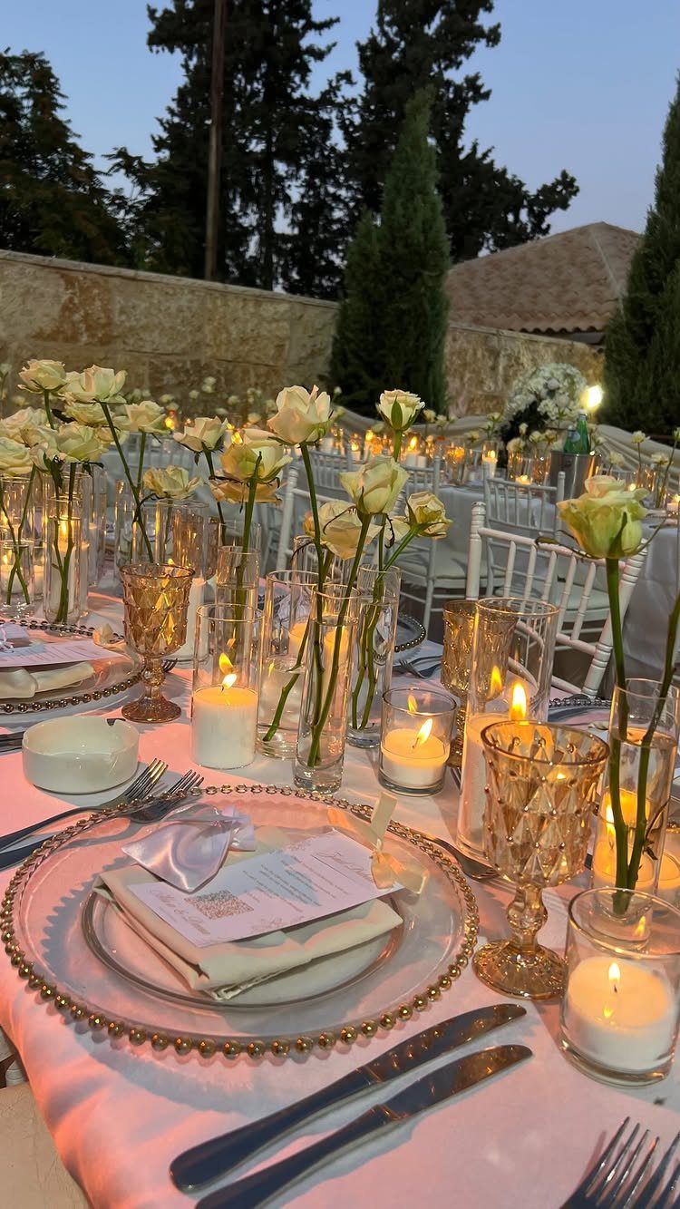 Candlelit wedding table with white roses, gold chargers, and glassware outdoors at dusk.