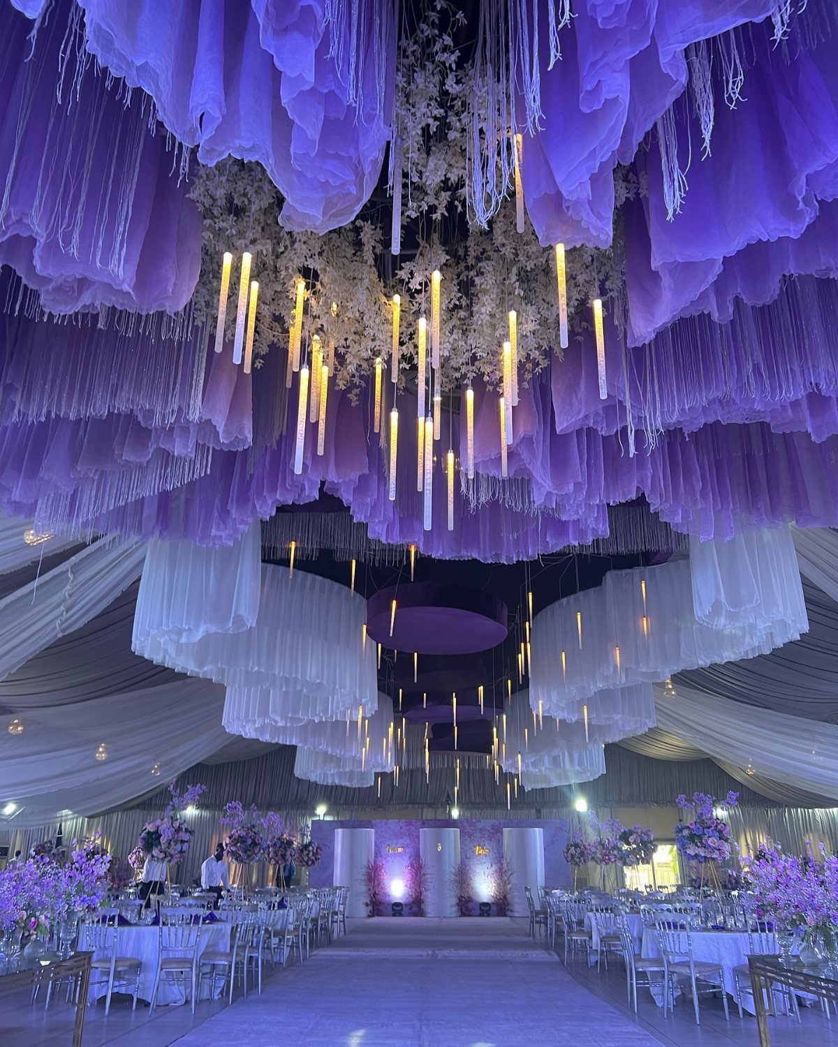 Lavish wedding reception hall with purple draped ceilings and hanging golden lights.