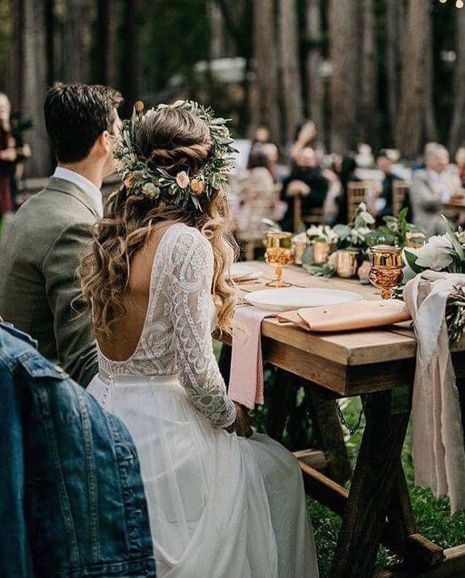 Bride with floral crown sitting at a rustic wooden table in a forest.