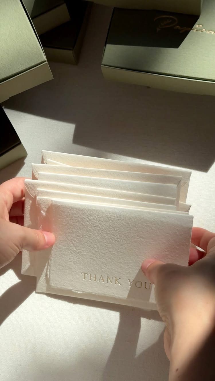 Hands holding a stack of white textured embossed thank you cards on a table.
