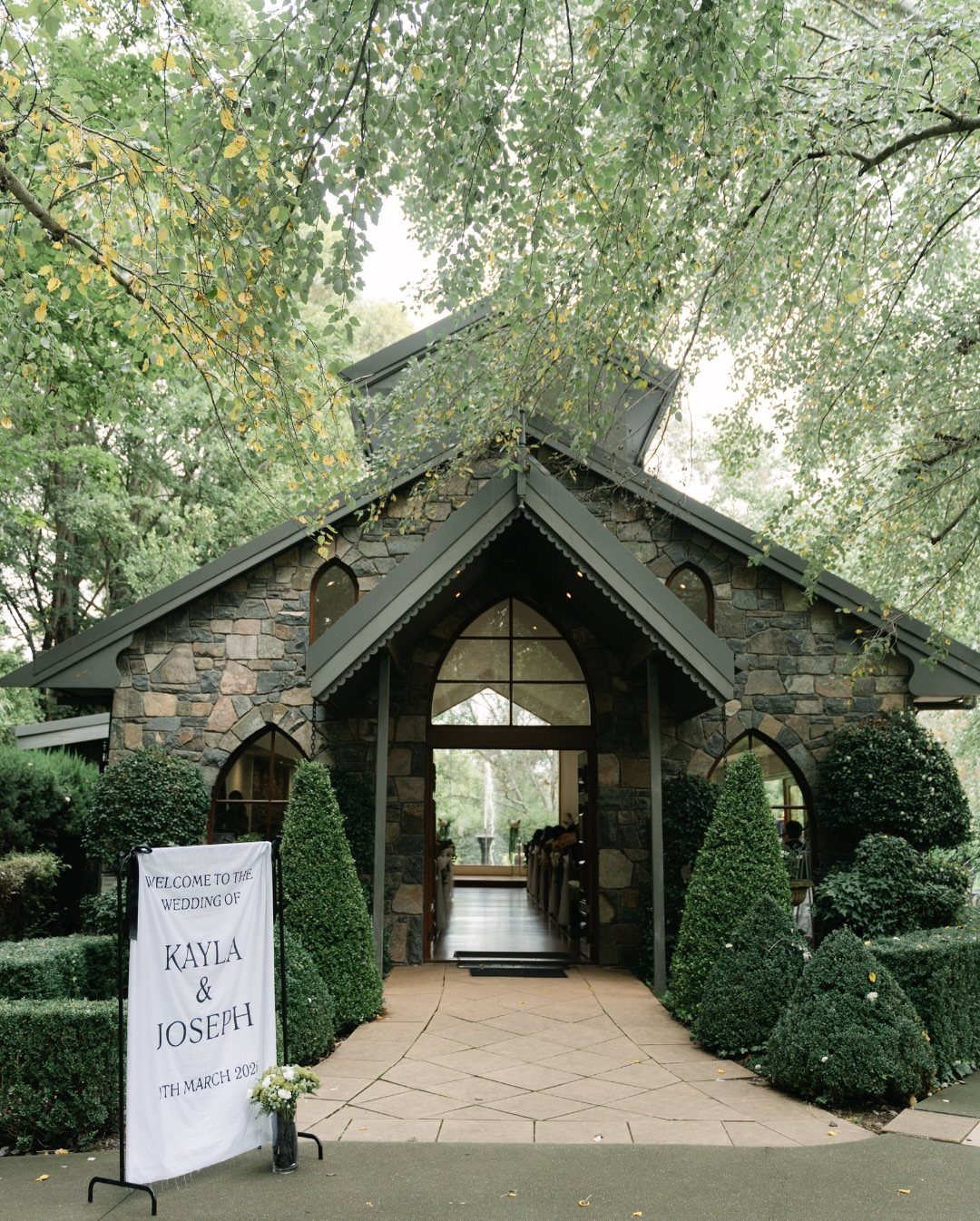 Stone chapel entrance with a wedding welcome sign and manicured green shrubbery.