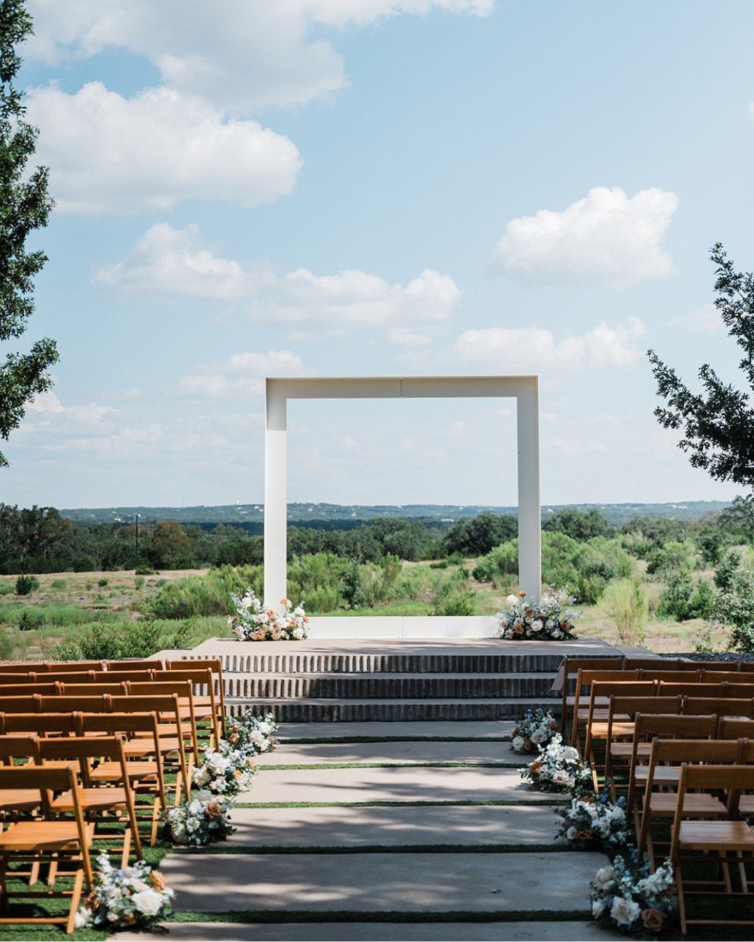 White rectangular wedding arch on a stone platform with wooden chairs and floral arrangements.