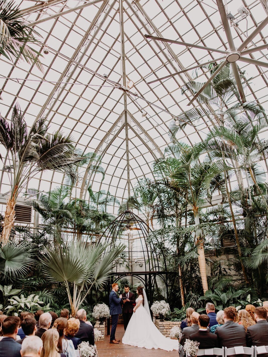 Wedding ceremony inside a glass conservatory filled with tropical palms and seated guests.