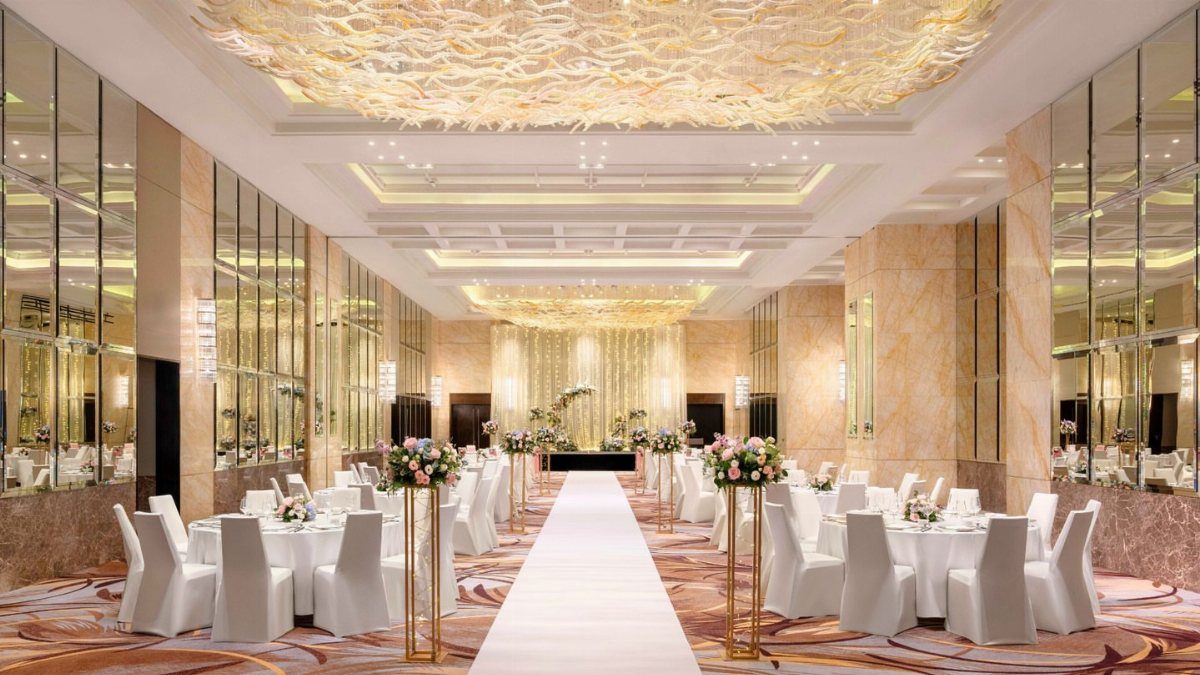 Luxurious hotel ballroom wedding setup with white tables, chairs, and gold ceiling decor.