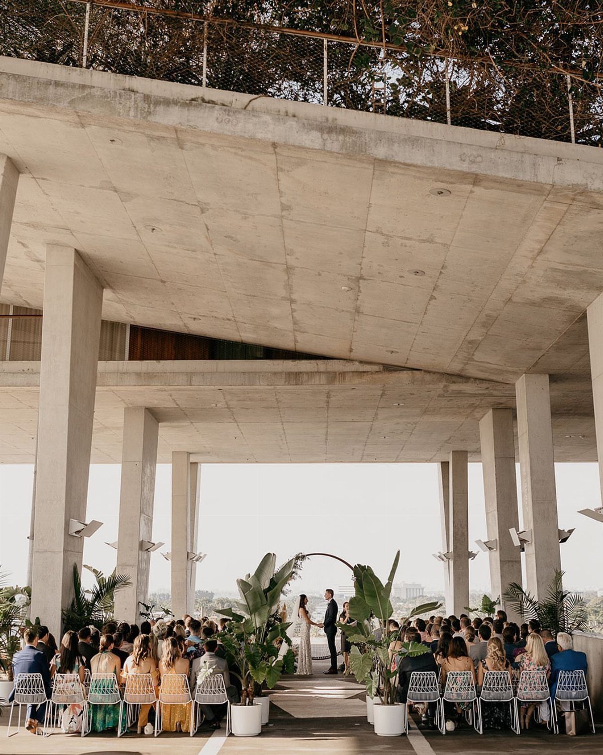 Wedding ceremony under a concrete structure with guests seated in white chairs.