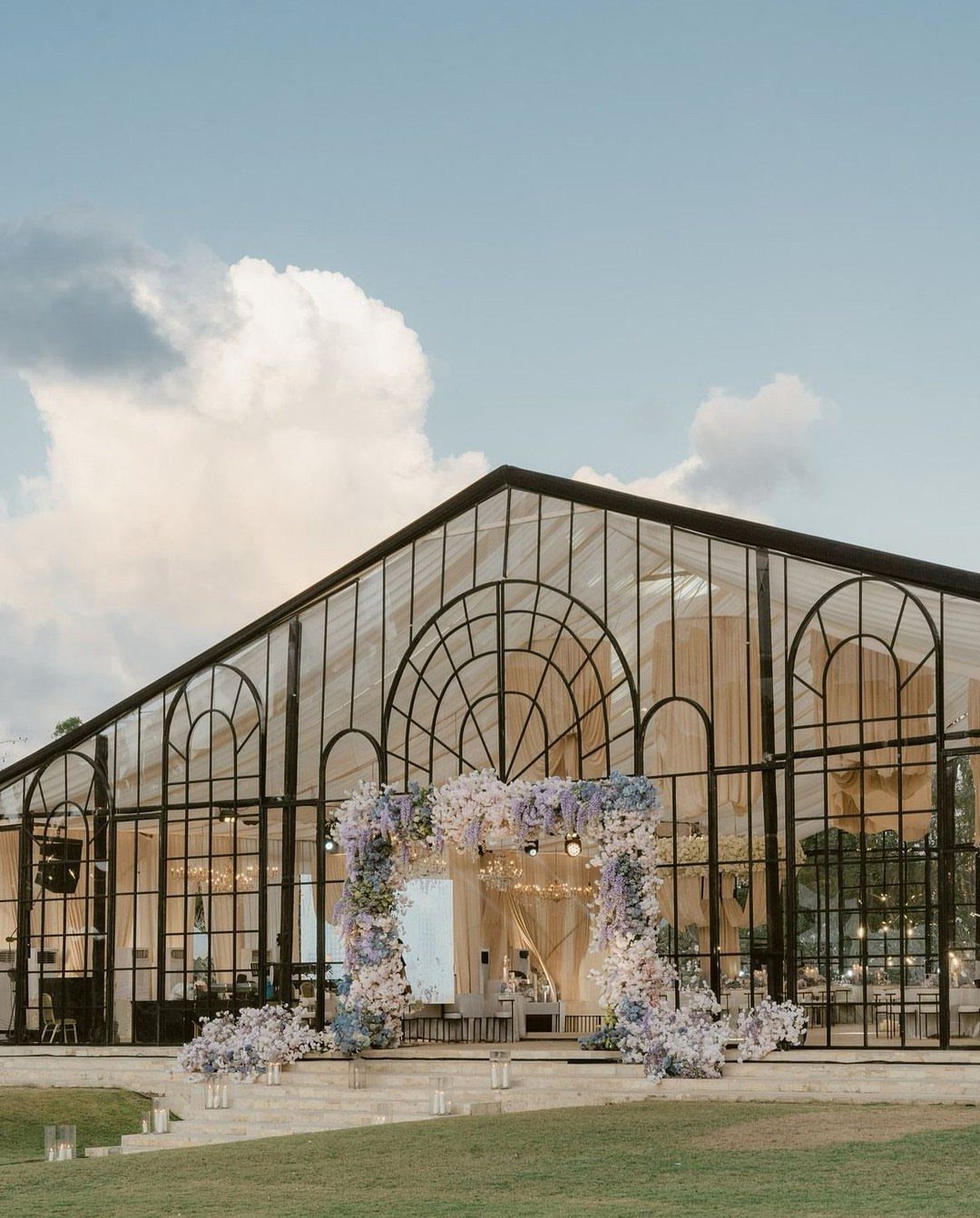 Modern glass greenhouse venue with black framing and a purple floral entrance arch.
