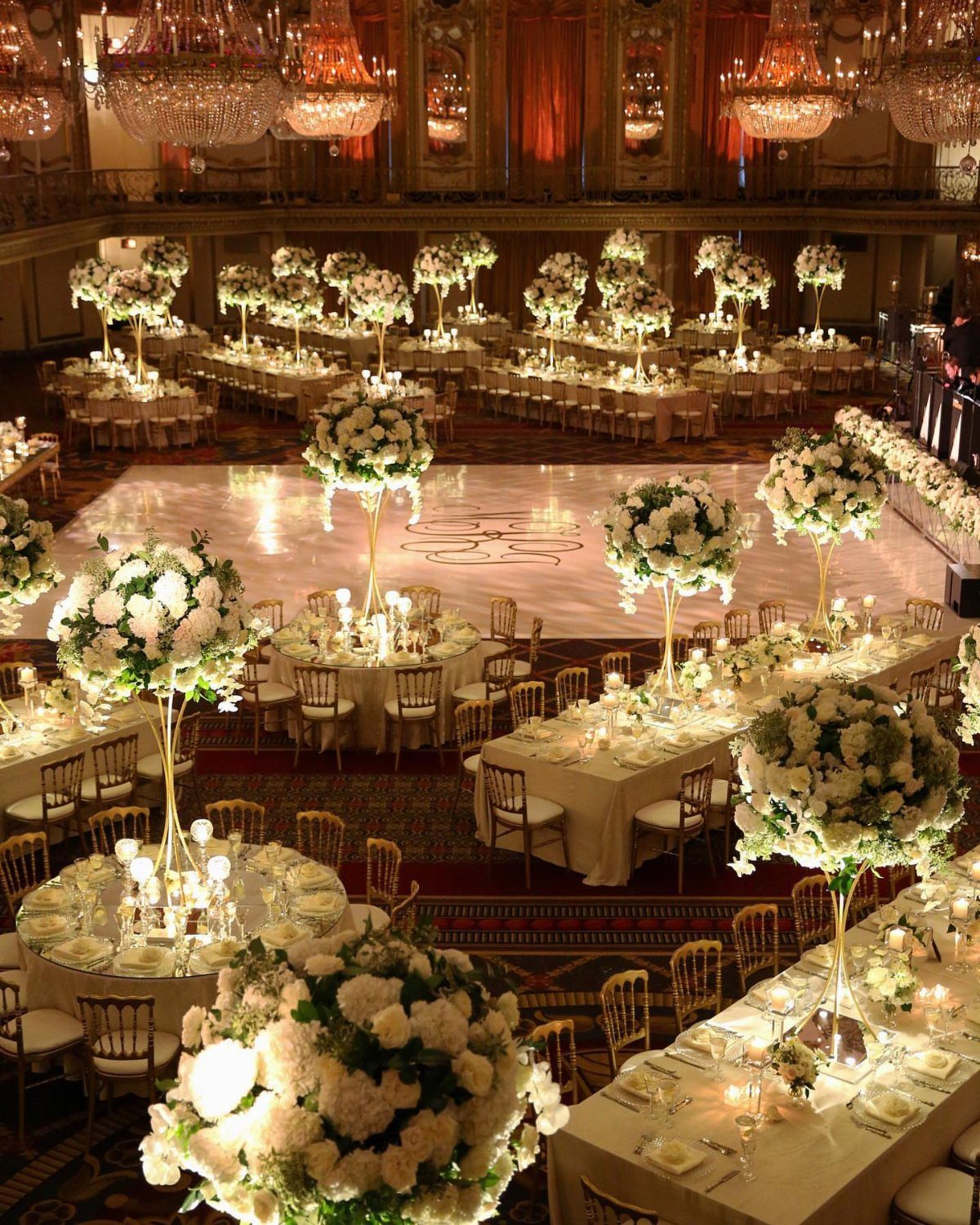 Opulent ballroom wedding reception with white floral centerpieces, gold chairs, and crystal chandeliers.