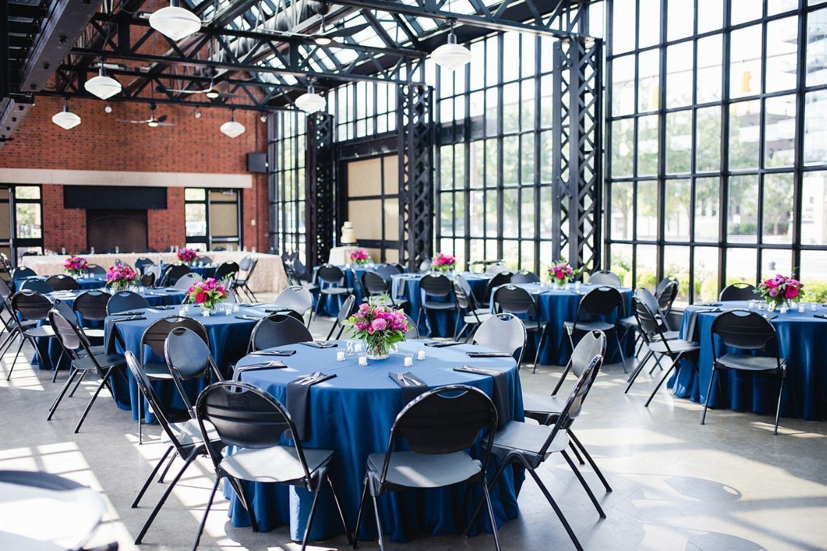 Industrial event space with blue clothed tables and pink flowers near large windows.