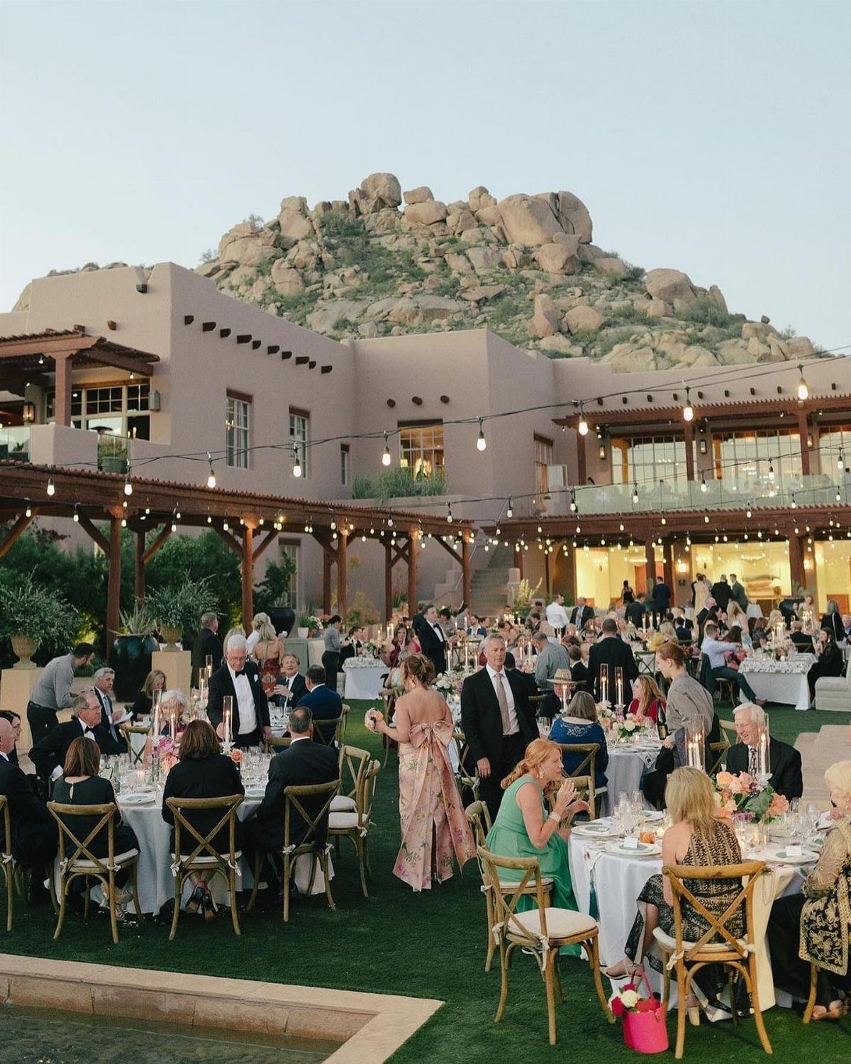 Outdoor wedding reception with guests at tables before a desert resort and rocky mountain.