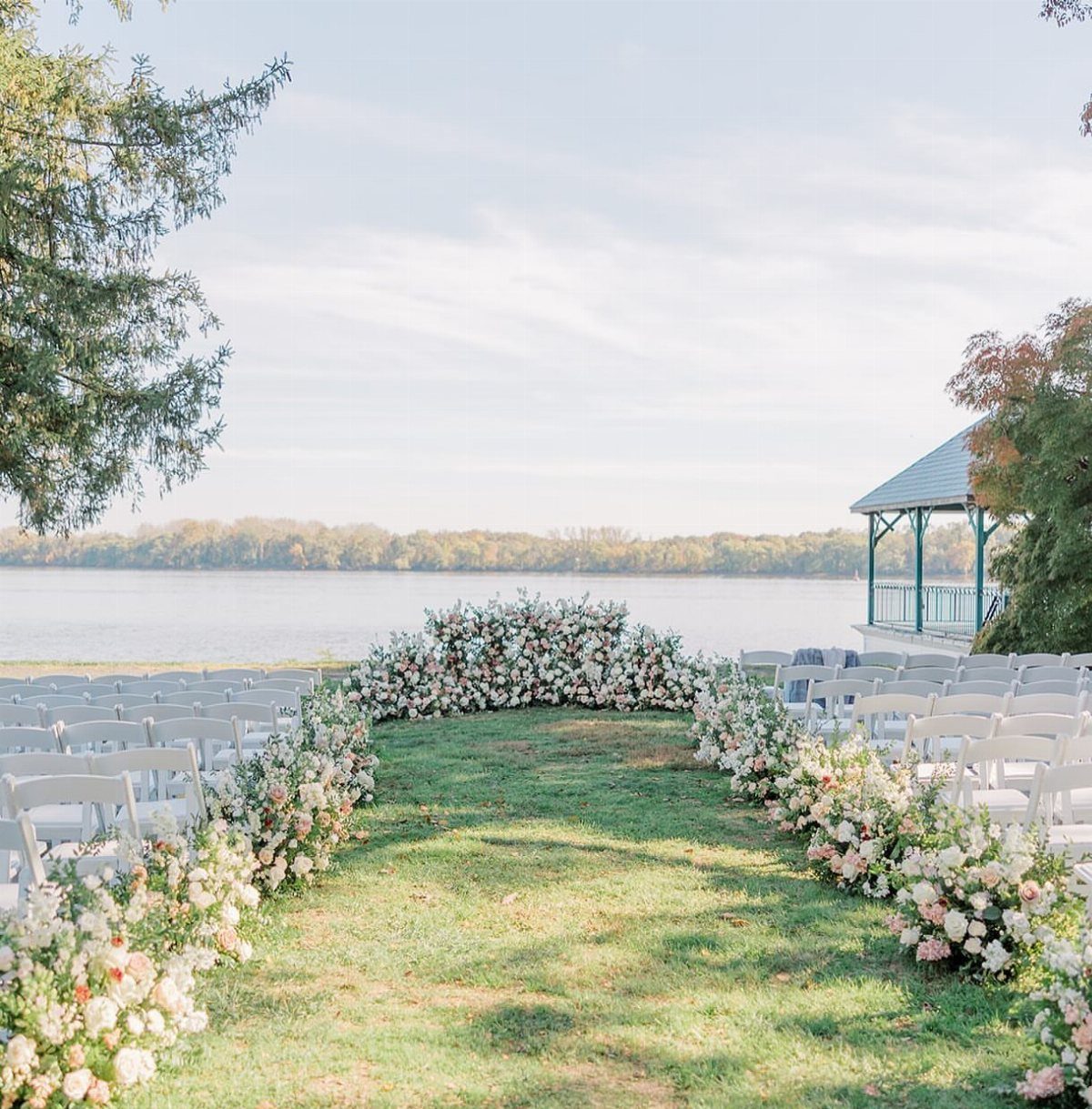 Outdoor wedding ceremony setup with white chairs and floral arch beside a calm lake.