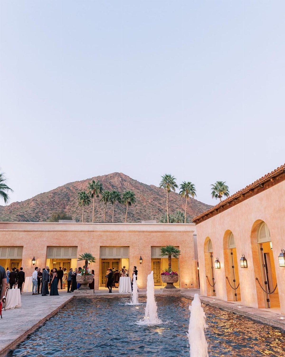 Mediterranean resort courtyard with fountains, formal guests, palm trees, and a mountain backdrop.