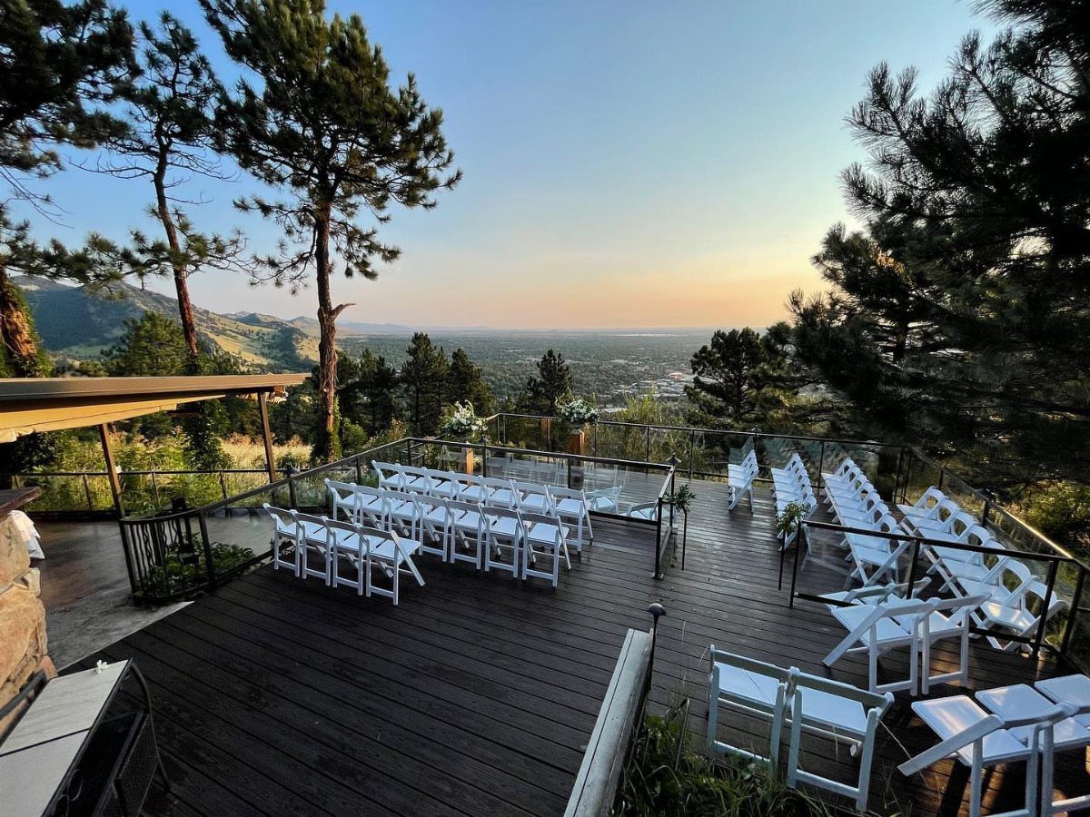 White wedding chairs on a wooden deck overlooking a mountain valley and pine trees.