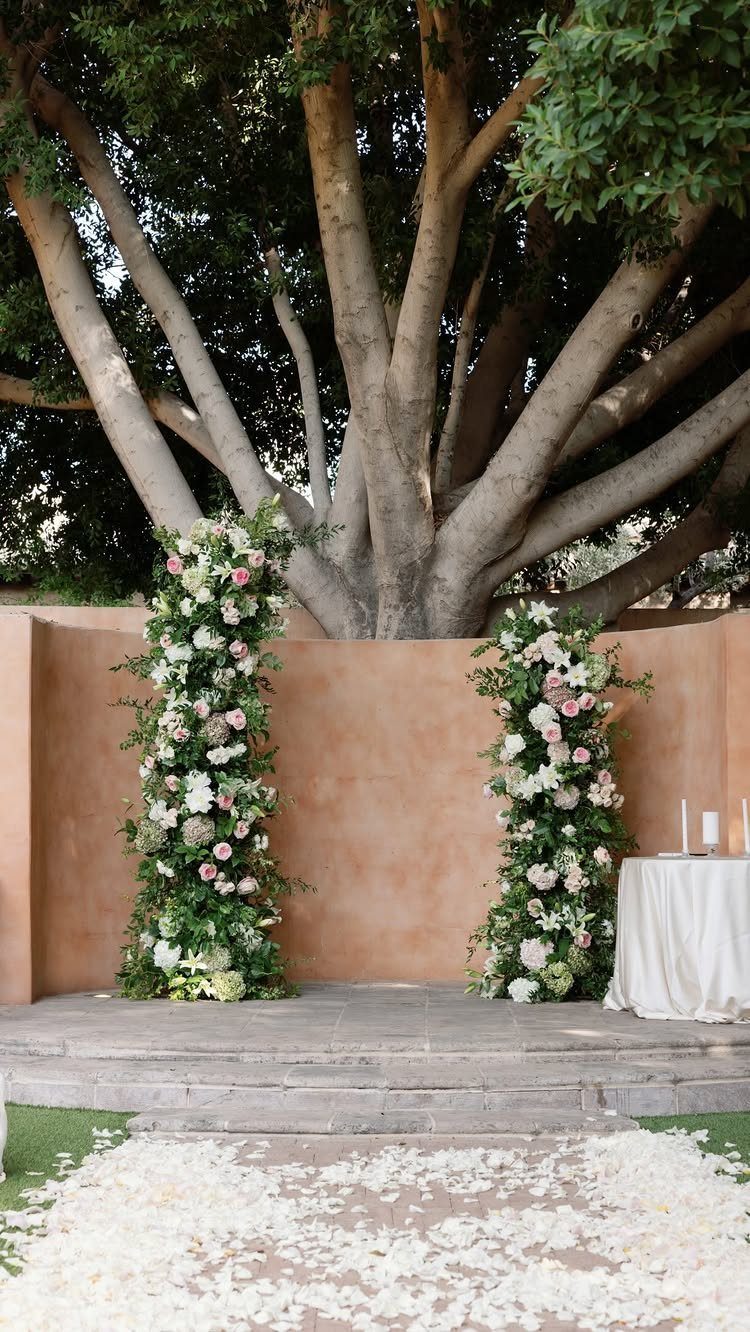 Wedding ceremony setup with floral pillars and a large tree against a terracotta wall.