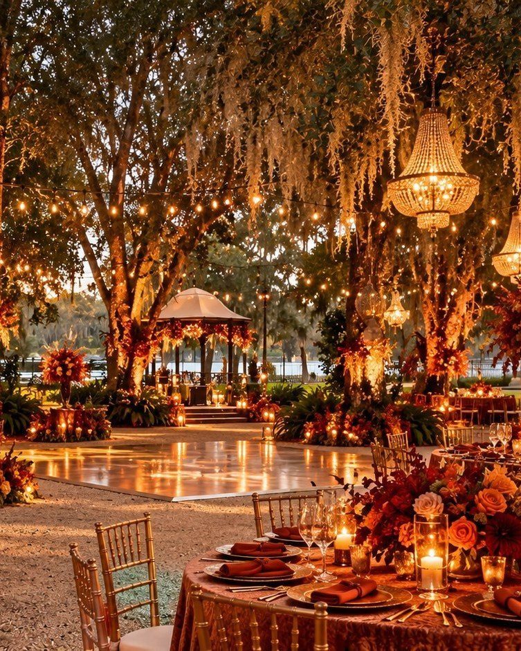 Outdoor wedding reception with chandeliers, string lights, and red flowers under mossy trees.