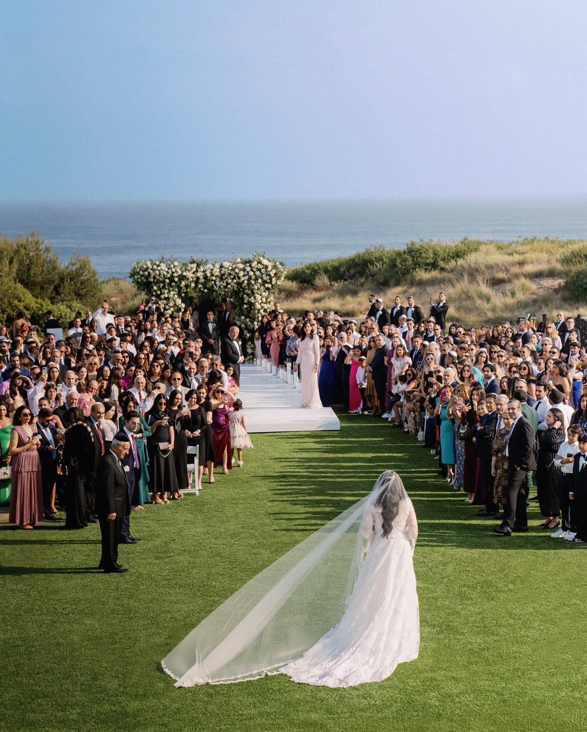 Bride walking toward a floral arch on a grassy lawn overlooking the ocean.
