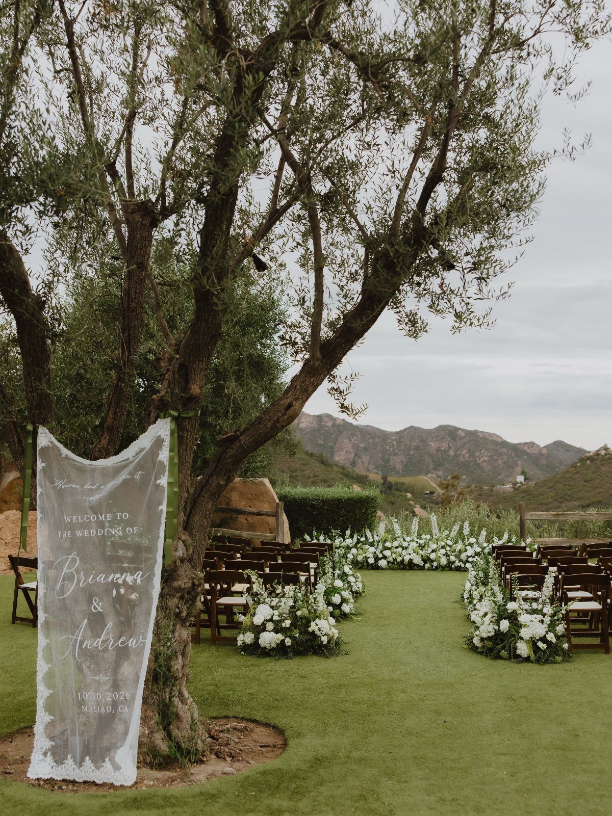 Outdoor wedding ceremony setup with wooden chairs, white flowers, and mountains in background.