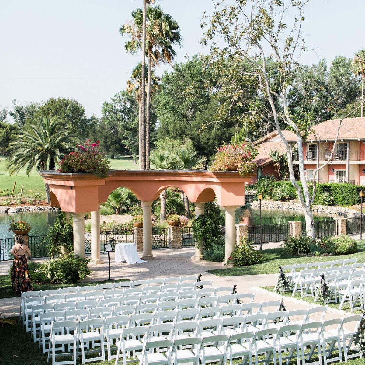 White wedding chairs facing a peach arched pavilion by a pond and palm trees.