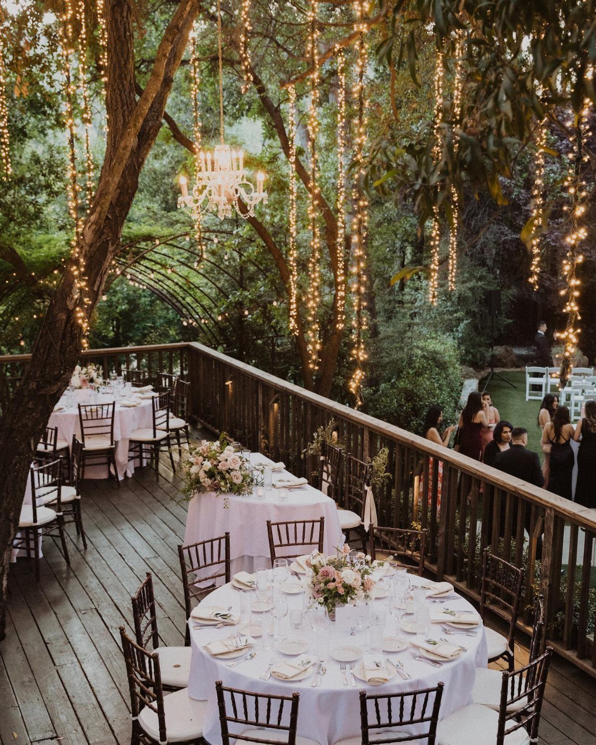Outdoor wedding reception on a wooden deck with string lights and white tables.