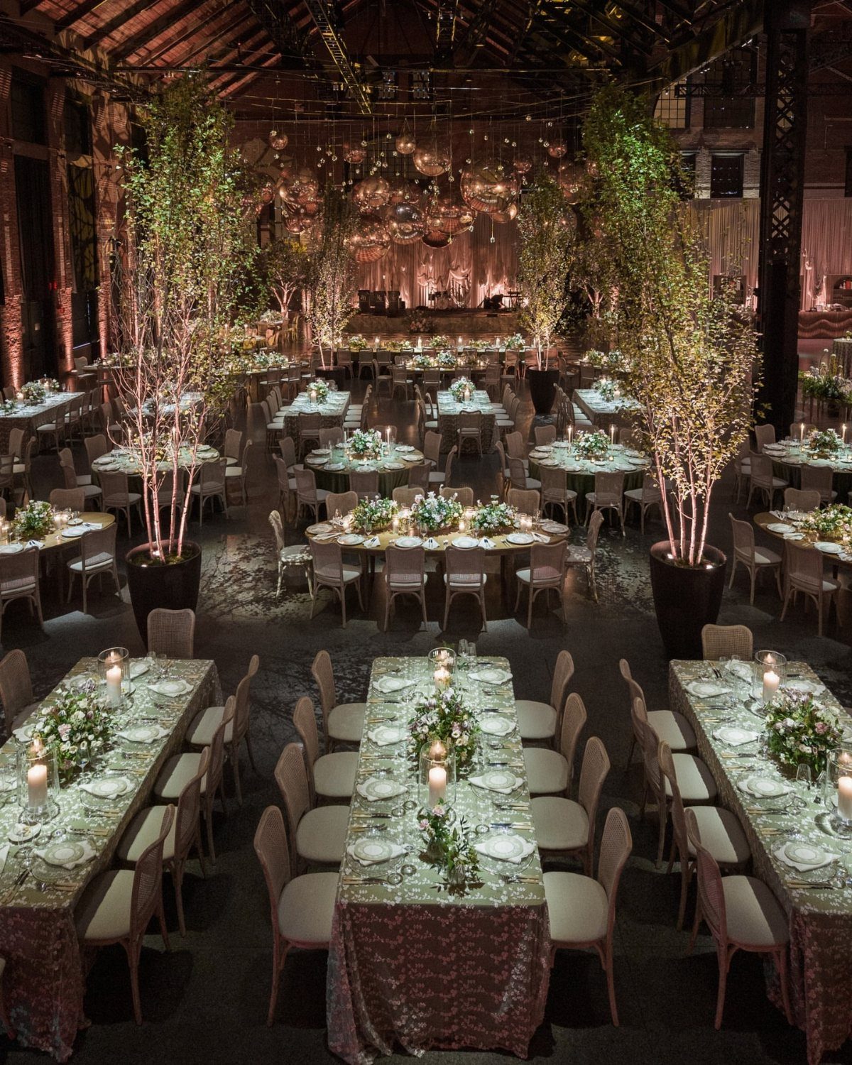 Industrial event space with dining tables, tall indoor trees, and hanging glass ornaments.