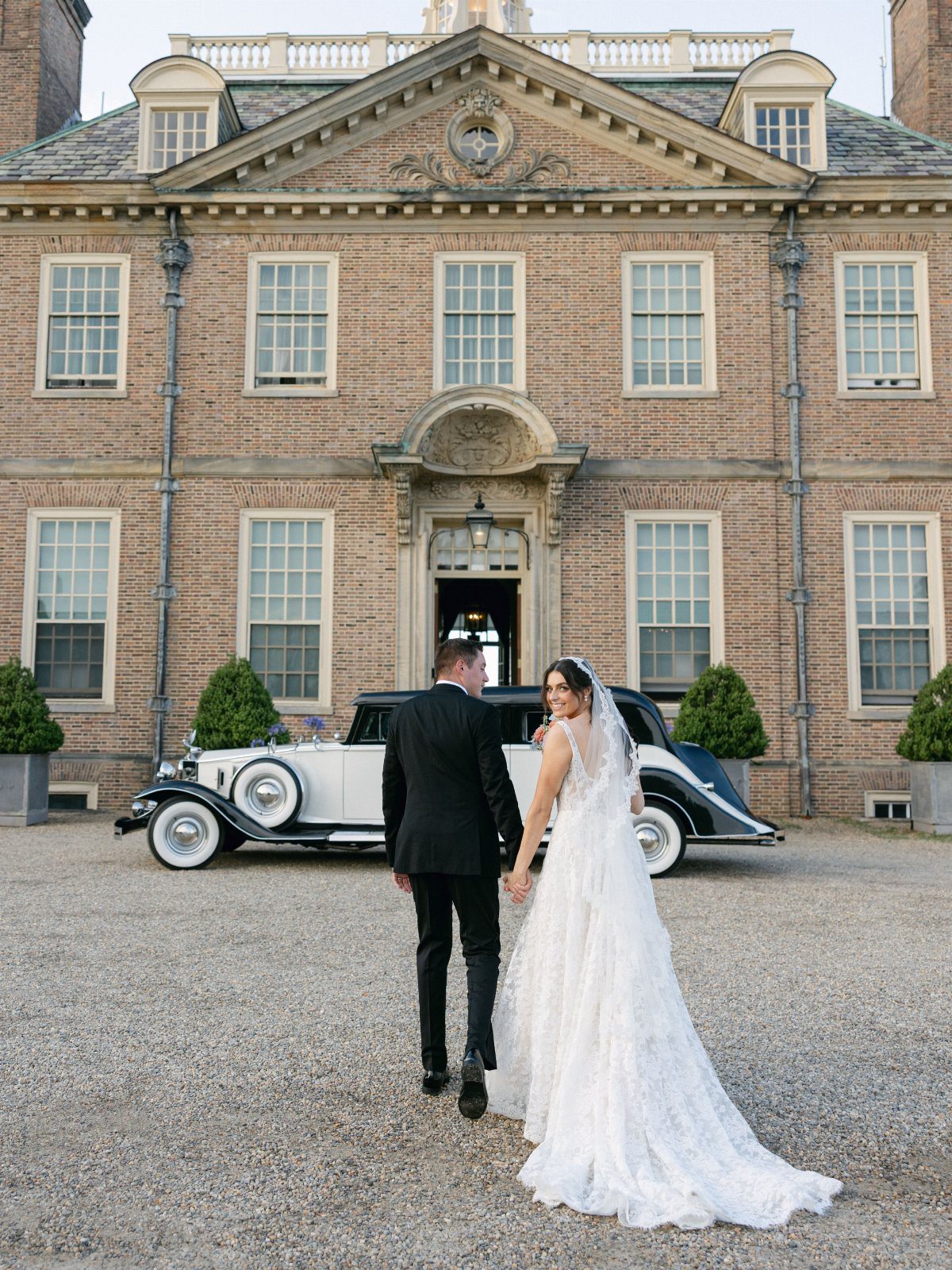 Bride and groom walking toward a historic brick mansion with a vintage car.