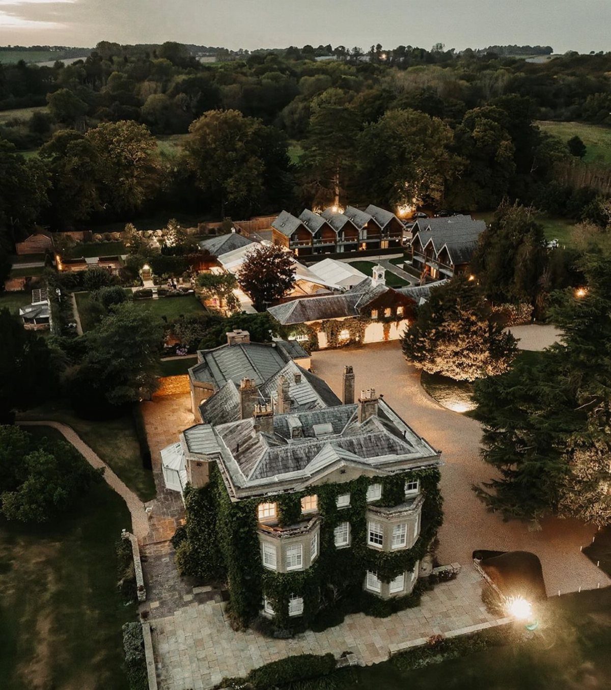 Ivy-covered historic stone mansion and estate buildings illuminated at dusk in a forest setting.
