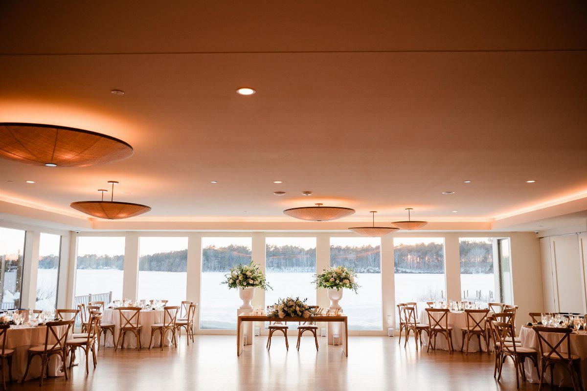 Modern waterfront wedding reception room with large windows overlooking a lake and forest.