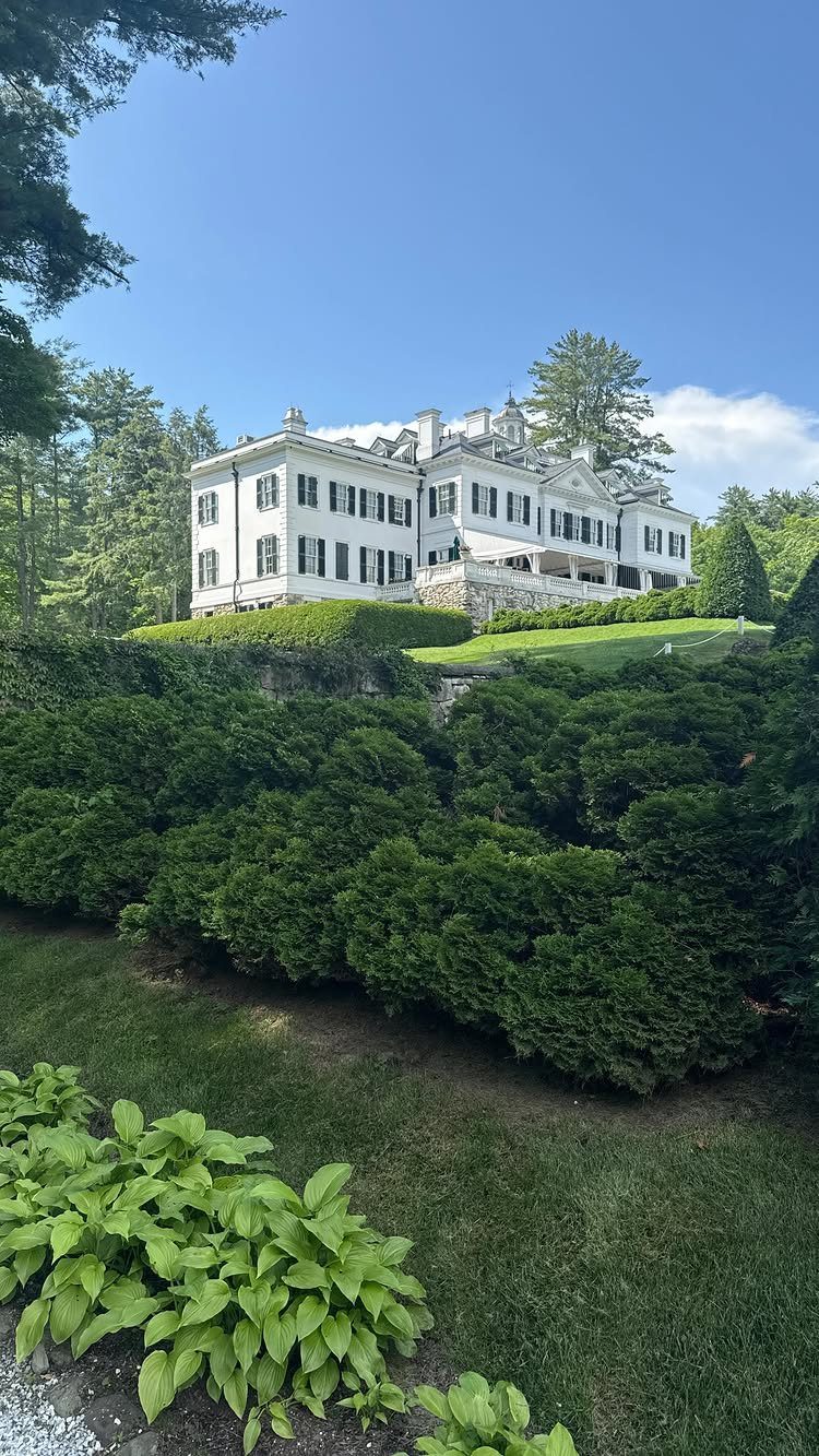 White historic mansion with black shutters overlooking lush green gardens and hedges.