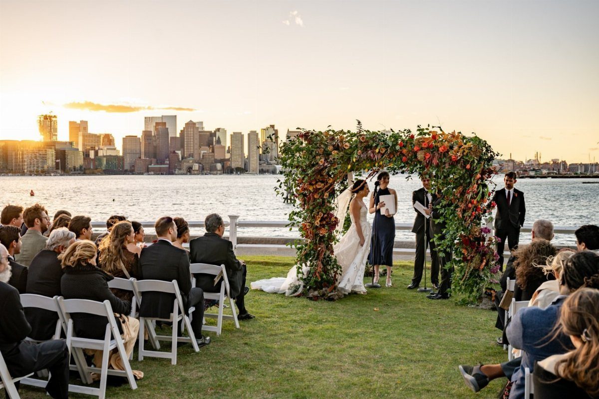 Wedding ceremony with a floral arch on a waterfront lawn overlooking a city skyline.
